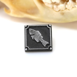 May include: A silver-colored fish design on a black square pin with a silver border and four silver studs.