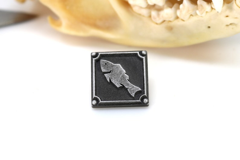 May include: A silver-colored fish design on a black square pin with a silver border and four silver studs.