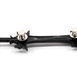 May include: Black plastic sword pin with silver metal pin backs. The sword has a detailed design with sharp points.