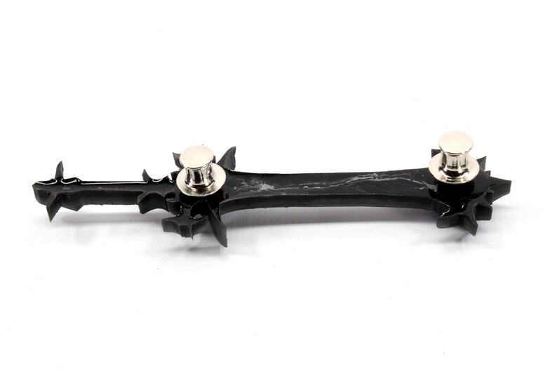 May include: Black plastic sword pin with silver metal pin backs. The sword has a detailed design with sharp points.