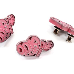 May include: Two pink heart-shaped pins with the text "DOK DOK CLUB" in white. The pins have a silver-colored metal backing with a pin post.