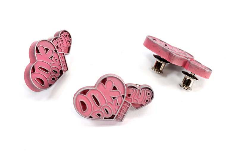 May include: Two pink heart-shaped pins with the text "DOK DOK CLUB" in white. The pins have a silver-colored metal backing with a pin post.