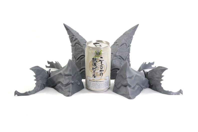 Op de afbeelding: Vijf 3D-geprinte grijze drakenkop-sculpturen rond een blikje Japans bier. Het blikje heeft Japanse tekst en de woorden "Japanese Craft Beer" erop gedrukt.