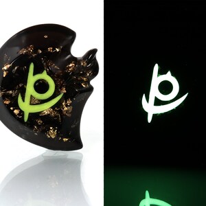 May include: A black resin pendant with gold flakes and a green glowing symbol. The symbol is a stylized letter "P" with a circle inside. The pendant glows green in the dark.