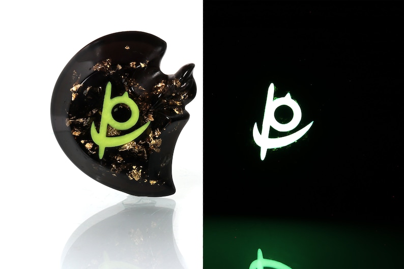 May include: A black resin pendant with gold flakes and a green glowing symbol. The symbol is a stylized letter "P" with a circle inside. The pendant glows green in the dark.