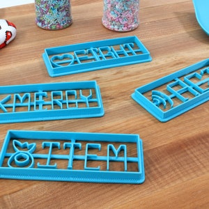 May include: Four blue plastic cookie cutters with different words and designs. The cutters spell out "Merry", "Heart", "Item", and "Sage".