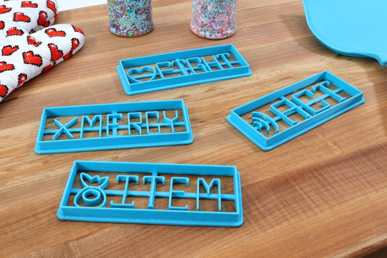 May include: Four blue plastic cookie cutters with different words and designs. The cutters spell out "Merry", "Heart", "Item", and "Sage".