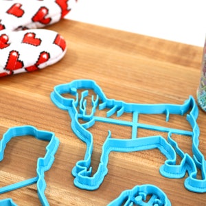 May include: A set of blue plastic dog-shaped cookie cutters on a wooden surface. A jar of colorful sprinkles is in the background.