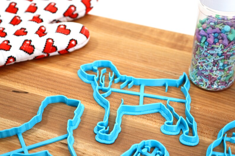 May include: A set of blue plastic dog-shaped cookie cutters on a wooden surface. A jar of colorful sprinkles is in the background.