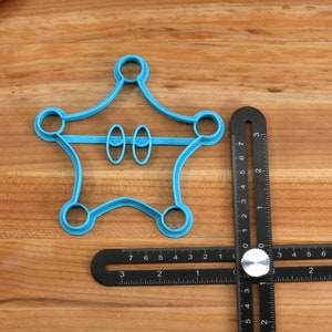 May include: A blue plastic cookie cutter shaped like a star with two circles in the middle. The cookie cutter is on a wooden surface with a black ruler.