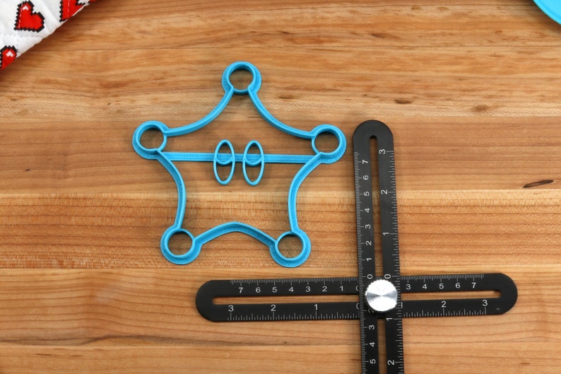 May include: A blue plastic cookie cutter shaped like a star with two circles in the middle. The cookie cutter is on a wooden surface with a black ruler.