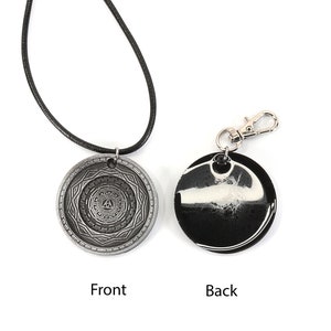 May include: A silver pendant necklace with a circular design featuring a geometric pattern and a triangle in the center. The back of the pendant is black with a white swirl design. The necklace is on a black cord and the pendant is also available as a keychain.