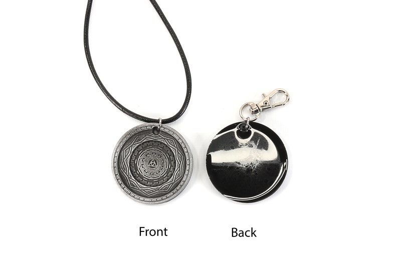 May include: A silver pendant necklace with a circular design featuring a geometric pattern and a triangle in the center. The back of the pendant is black with a white swirl design. The necklace is on a black cord and the pendant is also available as a keychain.