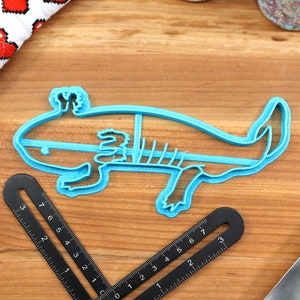 May include: A blue plastic cookie cutter shaped like an axolotl. The axolotl is a type of salamander that is native to Mexico.
