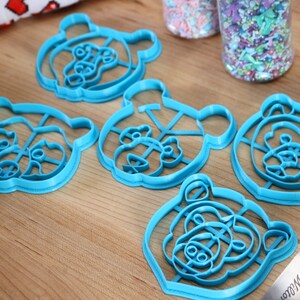 May include: Five blue plastic bear-shaped cookie cutters on a wooden surface.
