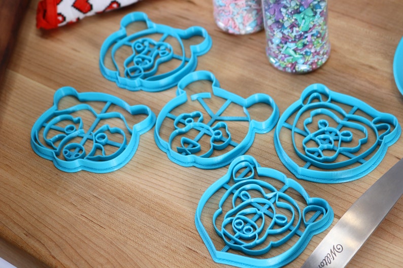 May include: Five blue plastic bear-shaped cookie cutters on a wooden surface.