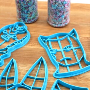 May include: Four blue plastic cookie cutters shaped like a cat, a dog, a snowman, and a Christmas tree. The cutters are on a wooden surface.