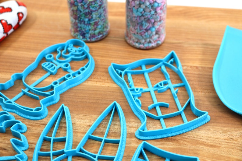 May include: Four blue plastic cookie cutters shaped like a cat, a dog, a snowman, and a Christmas tree. The cutters are on a wooden surface.