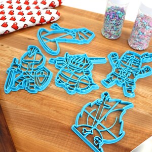 May include: A set of blue plastic cookie cutters shaped like knights and a ship. The cutters are on a wooden surface with sprinkles in jars and a rolling pin.