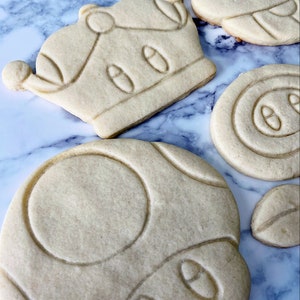 May include: A set of five sugar cookies decorated with royal icing. The cookies are shaped like a crown, a mushroom, and a star. The cookies are on a marble surface.