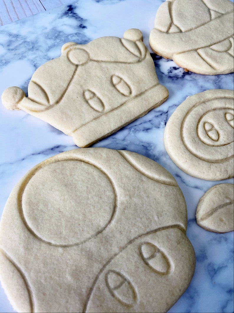 May include: A set of five sugar cookies decorated with royal icing. The cookies are shaped like a crown, a mushroom, and a star. The cookies are on a marble surface.