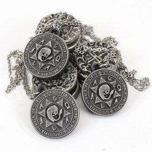 May include: Silver-toned pendant necklaces with intricate designs. Each circular pendant features a central emblem within a star, surrounded by text. The necklaces rest on a pile of similar, textured metal discs, all against a white background.