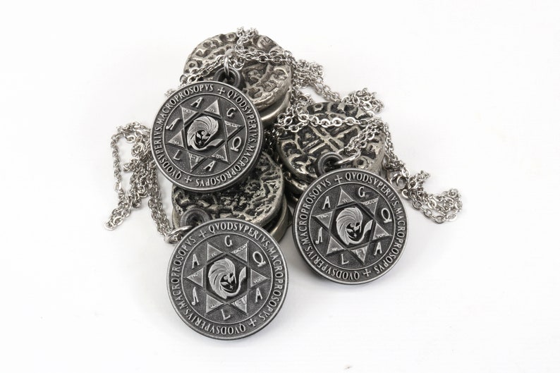 May include: Silver-toned pendant necklaces with intricate designs. Each circular pendant features a central emblem within a star, surrounded by text. The necklaces rest on a pile of similar, textured metal discs, all against a white background.