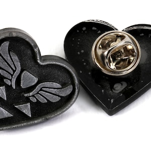 Puede incluir: Un pin de coraz&oacute;n negro con un dise&ntilde;o plateado de un coraz&oacute;n alado y tres tri&aacute;ngulos. La parte posterior del pin tiene un cierre de pin plateado.