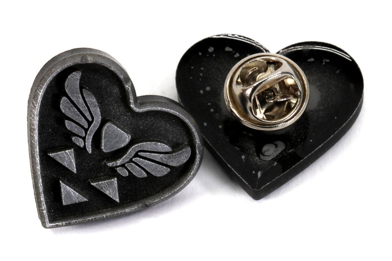Puede incluir: Un pin de coraz&oacute;n negro con un dise&ntilde;o plateado de un coraz&oacute;n alado y tres tri&aacute;ngulos. La parte posterior del pin tiene un cierre de pin plateado.