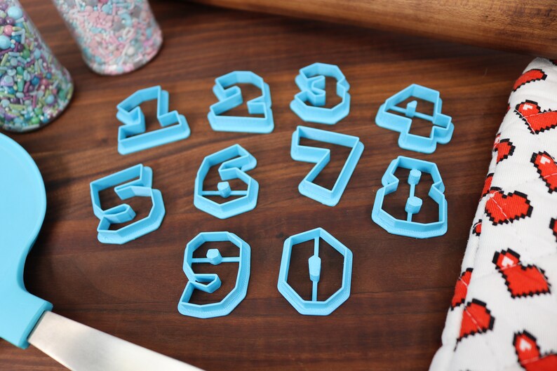 May include: A set of ten blue plastic cookie cutters shaped like the numbers 1 through 10. The cutters are on a wooden surface.