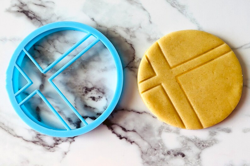 May include: A blue plastic cookie cutter shaped like a circle with four lines inside. A cookie is shown next to the cutter, with the same design imprinted on it.