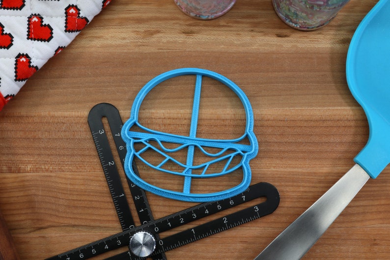 K&ouml;nnte beinhalten: Ein blauer Plastik-Ausstecher in Form eines Hamburgers. Der Ausstecher liegt auf einer Holzoberfl&auml;che mit einem Lineal und einem blauen Spatel.