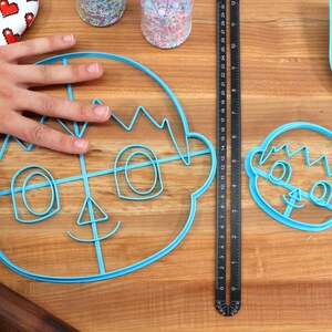 May include: Two blue plastic cookie cutters shaped like a cartoon face. The larger cutter has a grid pattern inside the face.