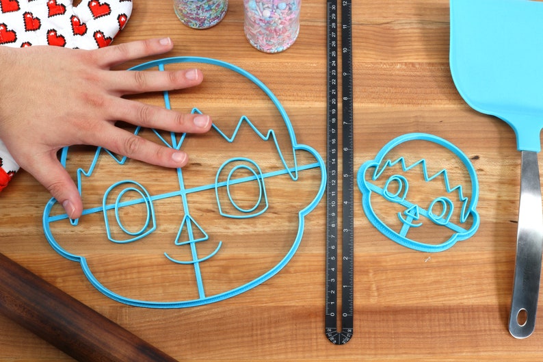 May include: Two blue plastic cookie cutters shaped like a cartoon face. The larger cutter has a grid pattern inside the face.