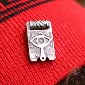May include: A silver-colored metal pin with a detailed design of a stylized eye with triangles and lines. The pin is on a red knitted beanie.