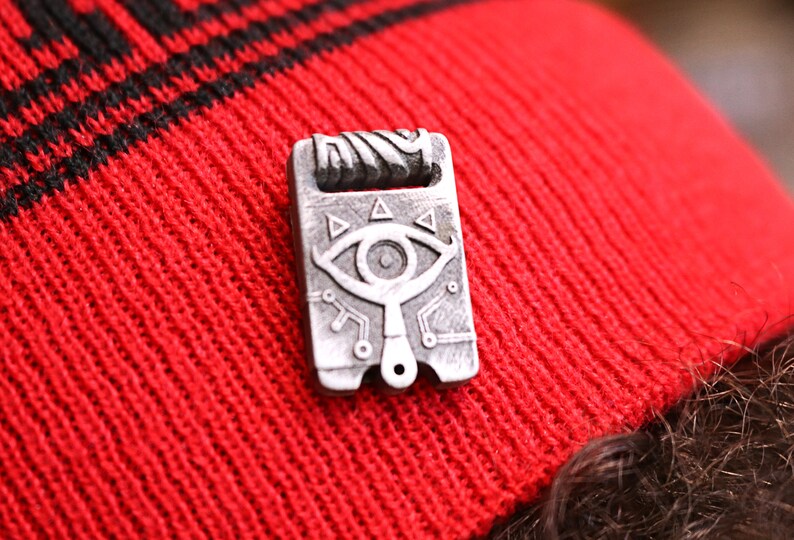 May include: A silver-colored metal pin with a detailed design of a stylized eye with triangles and lines. The pin is on a red knitted beanie.