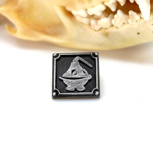 May include: A silver metal pin with a black background. The pin features a stylized image of a censer, a container used to burn incense.