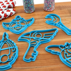 May include: A set of five blue plastic cookie cutters shaped like a crescent moon wand, a winged key, a winged heart, a winged shield, and a winged crown.