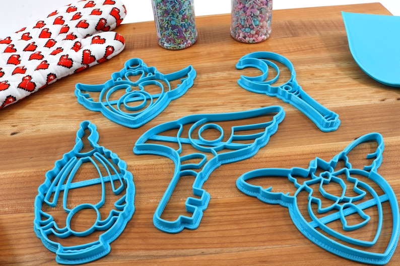 May include: A set of five blue plastic cookie cutters shaped like a crescent moon wand, a winged key, a winged heart, a winged shield, and a winged crown.
