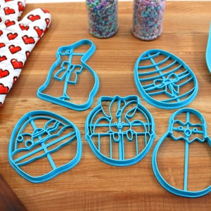 May include: A set of five blue plastic cookie cutters shaped like Easter eggs, bunnies, and chicks. The cutters are on a wooden surface with a rolling pin, a spatula, and two jars of sprinkles.