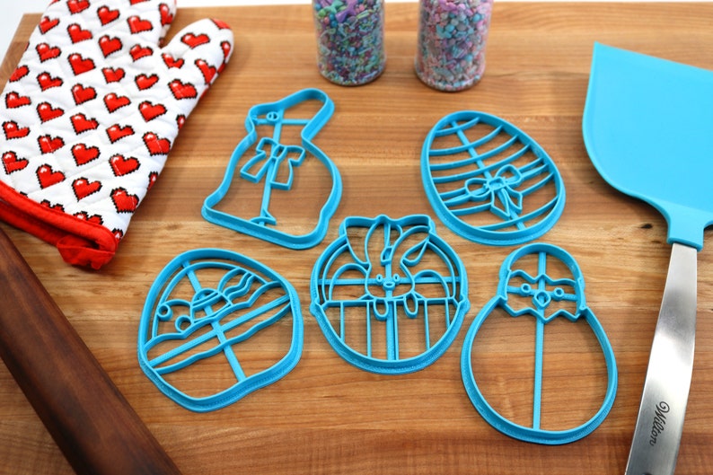 May include: A set of five blue plastic cookie cutters shaped like Easter eggs, bunnies, and chicks. The cutters are on a wooden surface with a rolling pin, a spatula, and two jars of sprinkles.