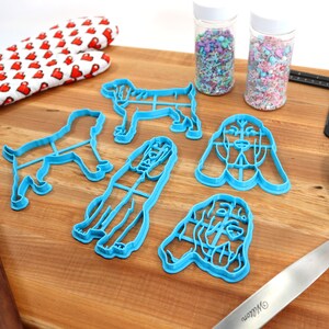 May include: A set of blue plastic dog-shaped cookie cutters on a wooden surface. There are also two jars of sprinkles, a rolling pin, a spatula, and a ruler.