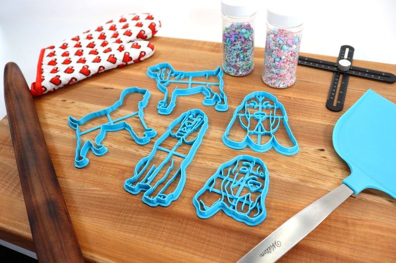 May include: A set of blue plastic dog-shaped cookie cutters on a wooden surface. There are also two jars of sprinkles, a rolling pin, a spatula, and a ruler.