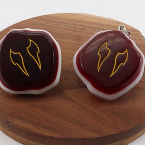 Op de afbeelding: Twee rode en witte hars cabochons met goudkleurige vlamontwerpen. De cabochons liggen op een houten oppervlak.