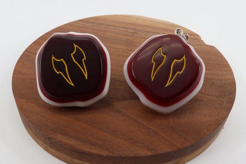 Op de afbeelding: Twee rode en witte hars cabochons met goudkleurige vlamontwerpen. De cabochons liggen op een houten oppervlak.