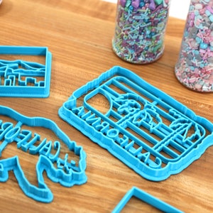 May include: Four blue plastic cookie cutters on a wooden surface. The cutters are shaped like a California palm tree, a bear with the word "California" written on it, a city skyline, and a rectangle.