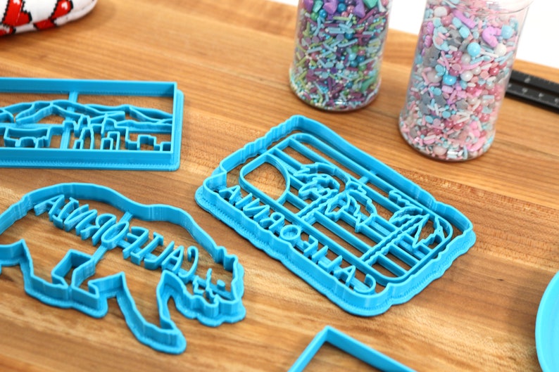 May include: Four blue plastic cookie cutters on a wooden surface. The cutters are shaped like a California palm tree, a bear with the word "California" written on it, a city skyline, and a rectangle.