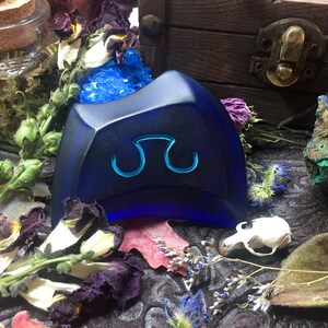 May include: A dark blue, triangular-shaped, glass paperweight with a stylized symbol in the center. The paperweight is sitting on a dark surface with dried flowers and other decorative items.
