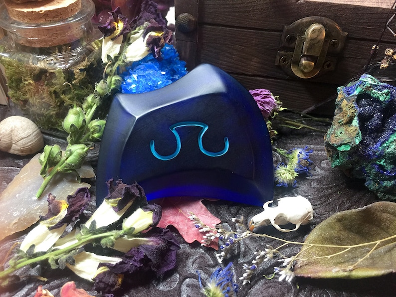 May include: A dark blue, triangular-shaped, glass paperweight with a stylized symbol in the center. The paperweight is sitting on a dark surface with dried flowers and other decorative items.