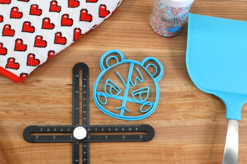 May include: Blue plastic cookie cutter shaped like a cartoon animal with a grumpy expression. The cutter is on a wooden surface with a ruler and a spatula.